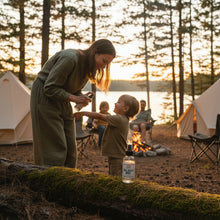 Load image into Gallery viewer, A clear glass spray bottle of Outdoor Bug Shield Spray with a black spray nozzle and a white label featured in an outdoor camp scene with mom, dad, and two kids, using the spray .
