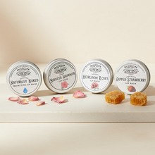 Load image into Gallery viewer, Four small round tins of lip balm arranged in a row on a rustic wooden surface, with a wooden plank background. The labels read Naturally Naked, Hibiscus Summer, Heirloom Roses, and Dipped Strawberry.