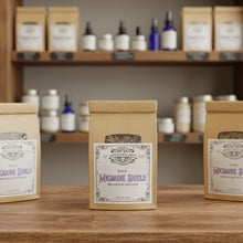 Load image into Gallery viewer, A kraft paper pouch labeled 'MacGuffin Shield' with a clear window showing dried herbs inside, displayed on a wood counter in a retail apothecary.