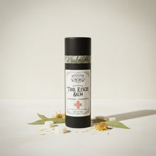 Load image into Gallery viewer, A tube of Herbal Trail Rescue Balm by The Healing Leaf Apothecary, lying on a light wood grain surface. The tube has a black cap, a white label with black text and a red cross, and a green patterned band near the cap.