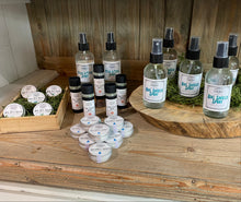 Load image into Gallery viewer, A collection of small personal care products arranged on a rustic wooden counter, including several clear spray bottles, several small black-and-white tins, and a wooden tray holding round white tins with labels. The products appear to be sold in the same style, with visible labels reading 'Bug Bite Balm', 'Trail Rescue Balm', Flea & Tick Spray', 'Naturally Naked Lip Balm', and 'Bug Shield Spray'