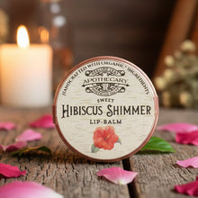 Load image into Gallery viewer, Lip balm with 'Sweet Hibiscus Shimmer' label on a wooden surface with pink petals and a candle in the background.