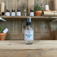 Load image into Gallery viewer, A clear glass spray bottle labeled 'Outdoor Bug Shield Spray' with a black spray top and white label, sitting on a rustic wooden surface against a wood-paneled background.
