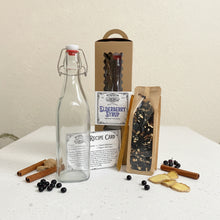 Load image into Gallery viewer, A glass swing-top bottle with a white stopper and metal clasp, standing beside a kraft paper box labeled Elderberry Syrup and a clear stand-up pouch filled with dark elderberry syrup and dried berries, all arranged on a textured beige tablecloth against a light wall.
