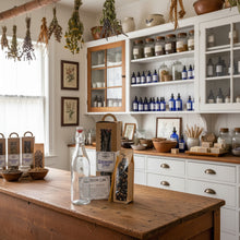 Load image into Gallery viewer, A glass swing-top bottle with a white stopper and metal clasp, standing beside a kraft paper box labeled Elderberry Syrup and a clear stand-up pouch filled with dark elderberry syrup and dried berries, all arranged in an apothecary setting
