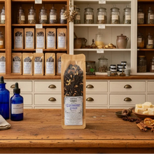 Load image into Gallery viewer, A kraft paper pouch of 'Exotic Spice Blend' herbal tea stands on a rustic wooden counter in a beautifully organized apothecary shop. The background features wooden and white shelves packed with glass jars of dried herbs, spices, and glass bottles, including cobalt blue bottles.
