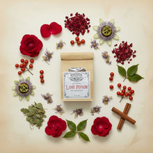 Load image into Gallery viewer, A kraft paper pouch labeled 'Love Potion' by Healing Leaf Apothecary styled with red rose petals, and small red berries, passionflowers, and cinnamon sticks on a light beige background.
