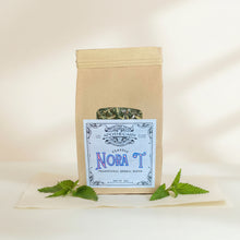Load image into Gallery viewer, A kraft paper pouch of Classic Nora Traditional Herbal Elixir by North Beauty Herb Co. displayed on a glass counter inside a shop, with the product label clearly visible.
