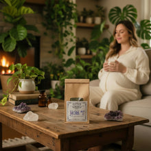 Load image into Gallery viewer, A kraft paper stand-up pouch of Classic Nora T Traditional Herbal Blend by Healing Leaf Apothecary, featuring a white label with blue and black text, set on a coffee table in front of a pregnant woman, by a fireplace and many green houseplants.

