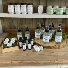 Load image into Gallery viewer, A display of small personal care or fragrance products arranged on a rustic wooden shelf, including several clear spray bottles, small dark bottles, round tins, and a tray of round lip balm or cream tins with white lids. The products have visible labels, and the setting includes moss and a wood slice prop.
