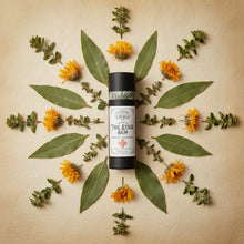Load image into Gallery viewer, A black cylindrical bottle of Herbal Trail Rescue Balm with a white label, styled on a light beige surface with green leaves, dried flowers, and white tablets.
