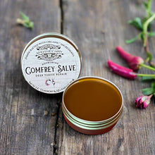 Load image into Gallery viewer, A small open tin container of Comfrey deep tissue repair salve placed on a wooden surface with comfrey leaves and cayenne peppers in the background.
