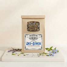 Load image into Gallery viewer, A kraft paper pouch labeled Apothecary Deep Stress Tonic stands on a white tray with scattered dried herbs and small blue flowers against a warm off-white background.

