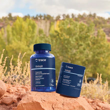 Load image into Gallery viewer, Two blue Trace Shilajit supplement bottles, one standing upright and one lying on its side, placed on a red rock outdoors with a blurred forest background.

